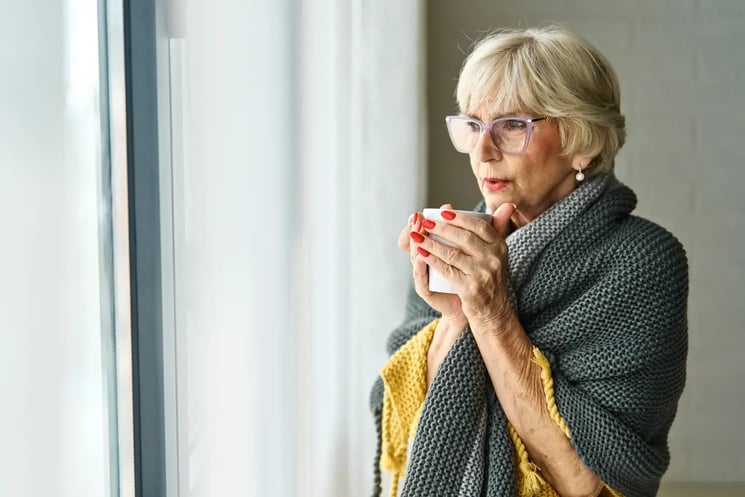 A woman with a blanket around her, drinking tea and looking out of the window